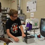 Two students are working together on a mechanical project in a classroom, with one guiding and the other operating equipment connected to a monitor displaying an enlarged view of their work.