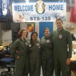 Four individuals in green jumpsuits stand together in a workplace setting under a banner reading "Welcome Home STS-135." Various flags and equipment are visible in the background.