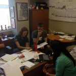 A group of seven people are seated around a table in an office, discussing and reviewing documents and notes. The room contains scientific posters, a whiteboard with graphs, and various items on shelves.