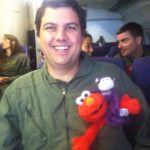 A man smiles on an airplane while holding a stuffed Elmo toy and a small purple monkey doll. Other passengers are visible in the background.