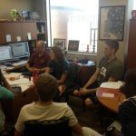 A group of people sit in an office around two desks with computers and papers, engaged in discussion.