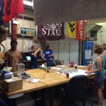 Several people are gathered around a large table in a workshop, working on various projects. Flags from different universities decorate the walls in the background.