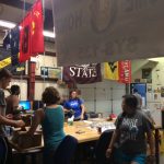 A group of people work together in a room with various state flags and equipment around. Some are seated at computers while others are standing and interacting near a table.