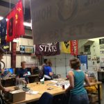 People working around a table in a room decorated with various university flags and maps, featuring shelves with equipment and posters on the walls.