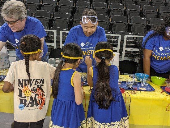Three children wearing yellow headbands and blue dresses participate in a science activity with two adult facilitators at a table covered in equipment and materials.