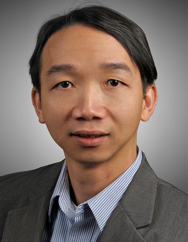A man with straight dark hair wearing a gray suit jacket and a blue striped shirt, facing the camera with a neutral expression against a plain background.