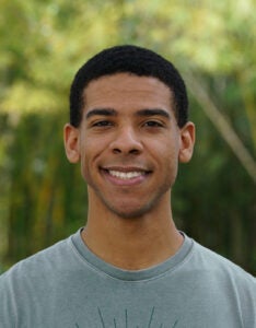 Smiling person wearing a green T-shirt with a circular design, standing outdoors with green foliage in the background.