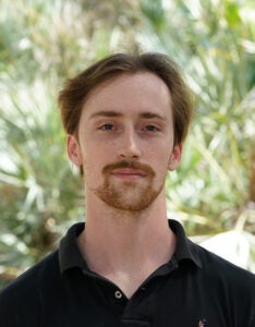 A person with short hair and a beard stands outdoors, wearing a black polo shirt. Greenery is blurred in the background.