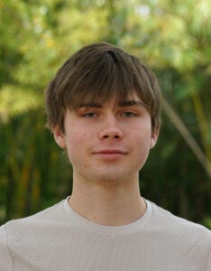 A young person with brown hair wearing a beige shirt stands outdoors with greenery in the background.