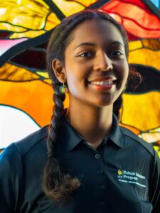 A person smiling in front of a colorful stained glass background, wearing a black McNair Scholars Program polo shirt.