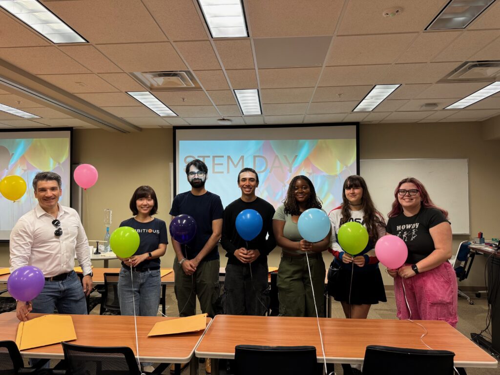 Seven people stand in a classroom holding balloons, with "STEM DAY" displayed on a screen behind them. Tables with folders and more balloons are in the foreground.
