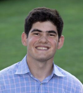 A young man with dark hair and thick eyebrows smiles at the camera, wearing a blue and white plaid button-up shirt, with a grassy background.