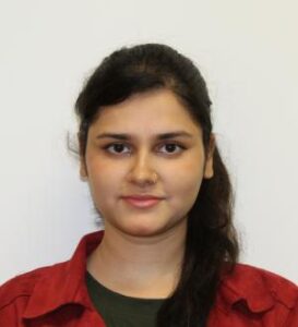A woman with long dark hair tied back, wearing a red collared shirt and dark top, is facing the camera against a plain white background.