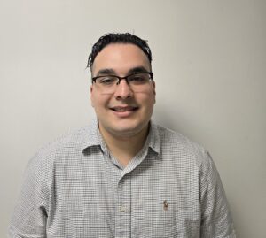 A man wearing glasses and a short-sleeve, checked button-up shirt stands smiling against a plain, light-colored wall.