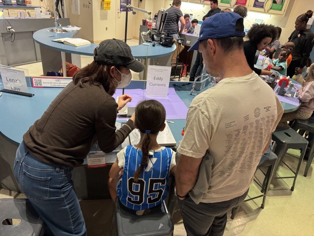 Three people, including a child in a blue sports jersey, are seated at a science exhibit table labeled "Eddy Currents" in a busy classroom or museum setting.