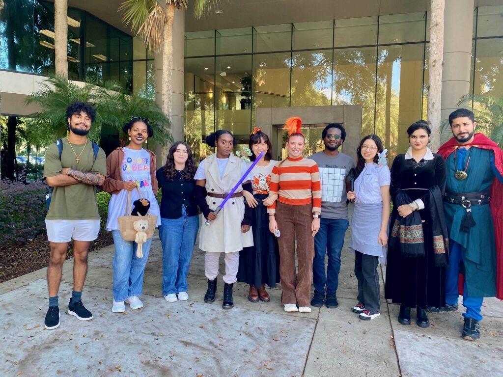 A group of ten people stand in a row outside a building; several are dressed in costumes, while others wear casual clothing.