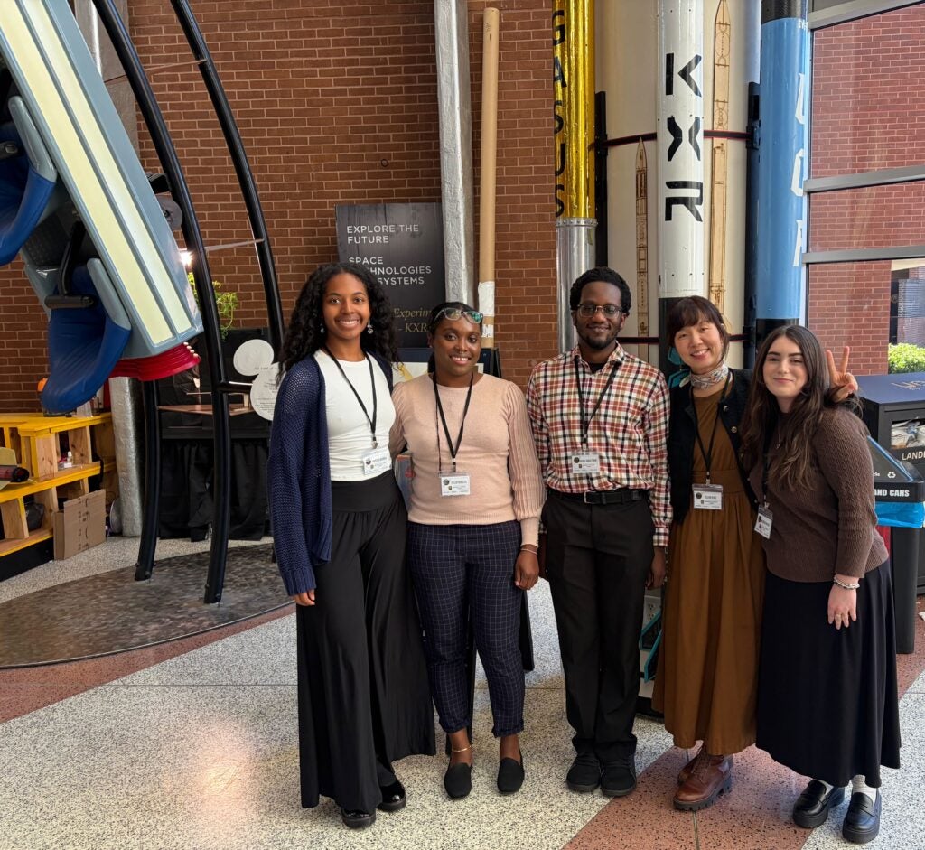 Five people stand indoors in front of large model rockets, smiling at the camera. All are wearing name badges and business casual clothing.