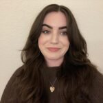 A woman with long brown hair wearing a brown sweater and a heart-shaped necklace is standing in front of a light-colored textured wall, smiling softly at the camera.