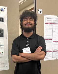 A person with curly hair stands smiling with arms crossed in front of a research poster display, wearing a black polo shirt and a name tag.
