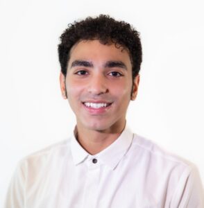 A person with short curly hair wearing a white button-up shirt, smiling and facing the camera against a plain white background.