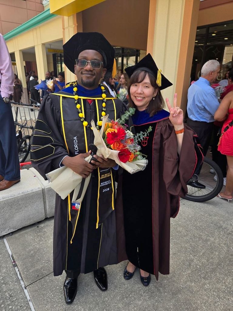 Two graduates in caps and gowns stand side by side, holding bouquets and smiling at the camera, with people and a building in the background.