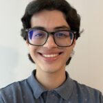Young person with dark hair and glasses smiling at the camera, wearing a blue collared shirt with small white prints, standing against a plain light-colored wall.