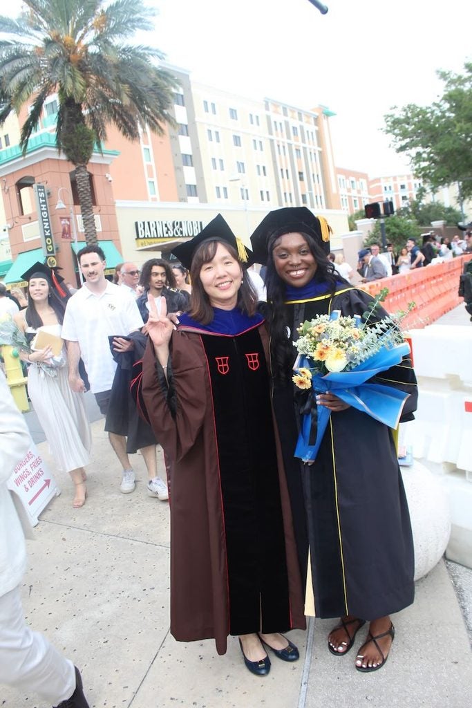 Two women in academic regalia stand outdoors, one holding a bouquet of flowers, smiling at the camera after a graduation ceremony. People and buildings are visible in the background.