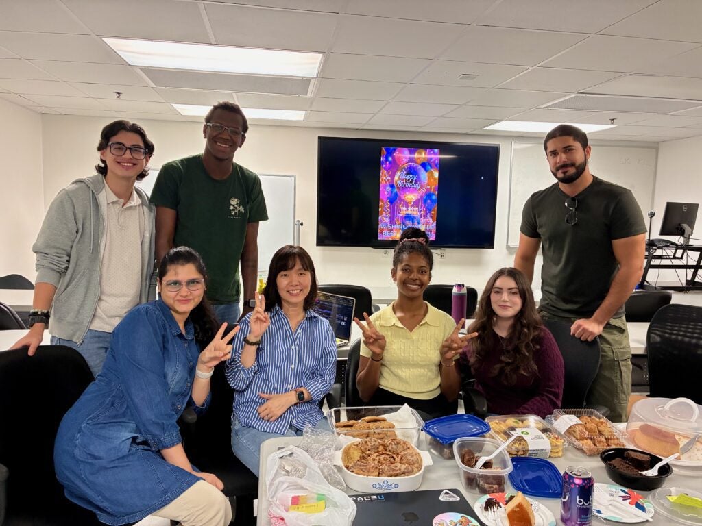 Seven people pose for a group photo in an office breakroom with a birthday message displayed on a monitor in the background.