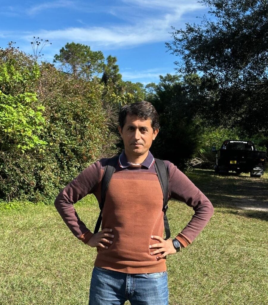 A man wearing a brown sweater and backpack stands with hands on hips on a grassy area, with trees and a vehicle in the background under a blue sky.