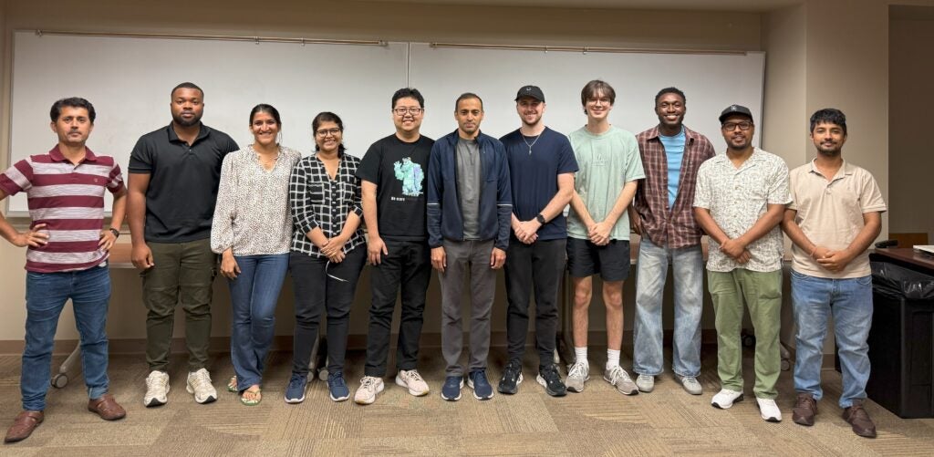 A group of eleven people stand side by side in a classroom with whiteboards in the background, posing for a photo.