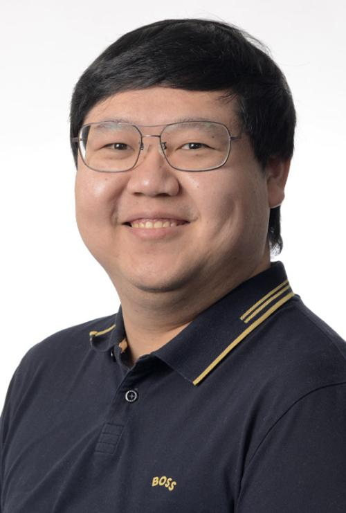 A person with short black hair and glasses is smiling at the camera, wearing a black collared shirt with yellow trim.