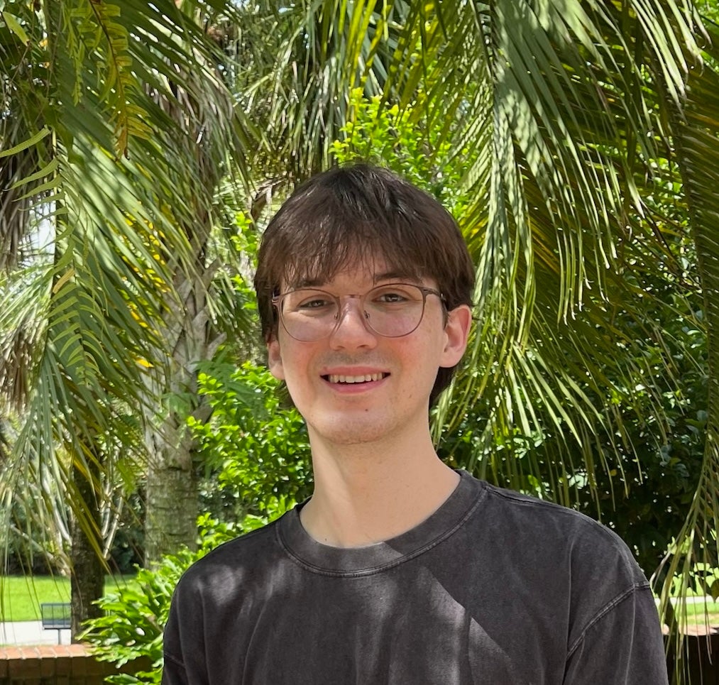 A young person with glasses and brown hair smiles outdoors, standing in front of green palm leaves and sunlight.