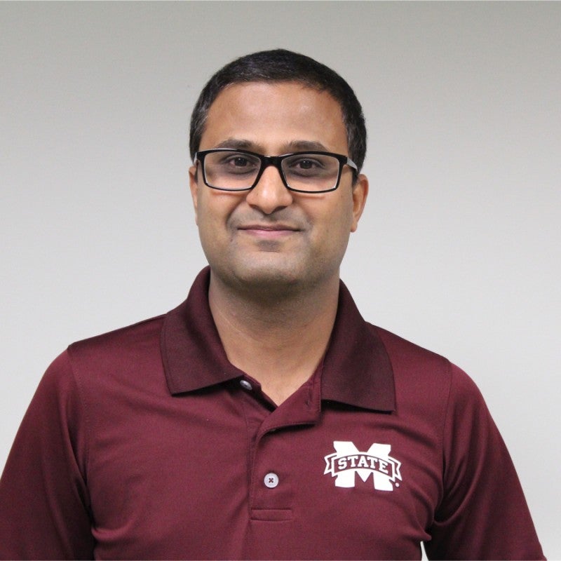A man with short dark hair and glasses wearing a maroon Mississippi State University polo shirt stands in front of a plain light-colored background.