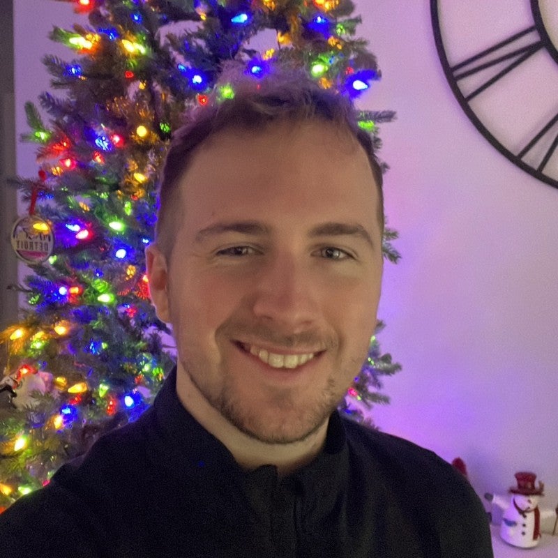 A person smiling in front of a decorated Christmas tree with colorful lights; a wall clock and a small snowman decoration are visible in the background.