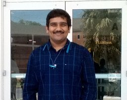 A person with short dark hair and a mustache, wearing a blue checkered shirt, stands smiling in front of glass doors.