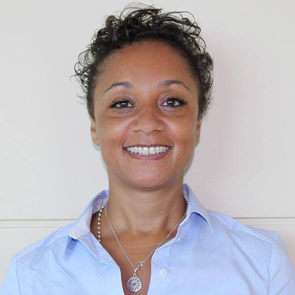 A person with curly hair is smiling and wearing a blue button-up shirt and a necklace with a circular pendant against a plain background.