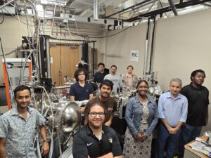 A group of nine people stand and smile in a laboratory filled with scientific equipment, wires, and machinery.