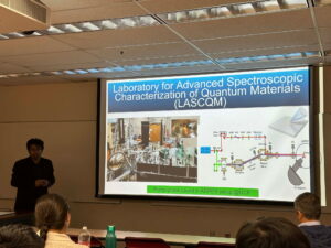 A presenter stands beside a screen displaying information and diagrams about the Laboratory for Advanced Spectroscopic Characterization of Quantum Materials at UCF in a classroom setting.