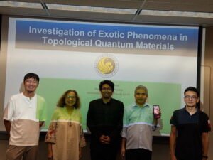 Five people stand in front of a presentation slide titled "Investigation of Exotic Phenomena in Topological Quantum Materials"; one person holds a phone showing another person's face.