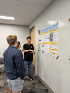 Three people stand indoors as one presents research on a wall-mounted poster. The presenter gestures toward the poster while the others listen attentively.