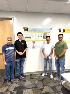 Four men stand indoors in front of a scientific research poster taped to a wall. All are facing the camera. The floor has a geometric carpet pattern.