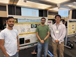 Three men stand in front of a scientific research poster at a conference, with more posters and attendees visible in the background.