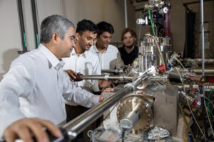 Four people observe and adjust a complex piece of scientific equipment with metal tubes, wires, and gauges in a laboratory setting.
