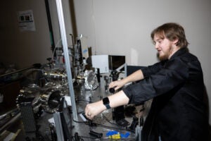 A person adjusts equipment on an optical table in a laboratory, surrounded by scientific instruments, cables, and tools.