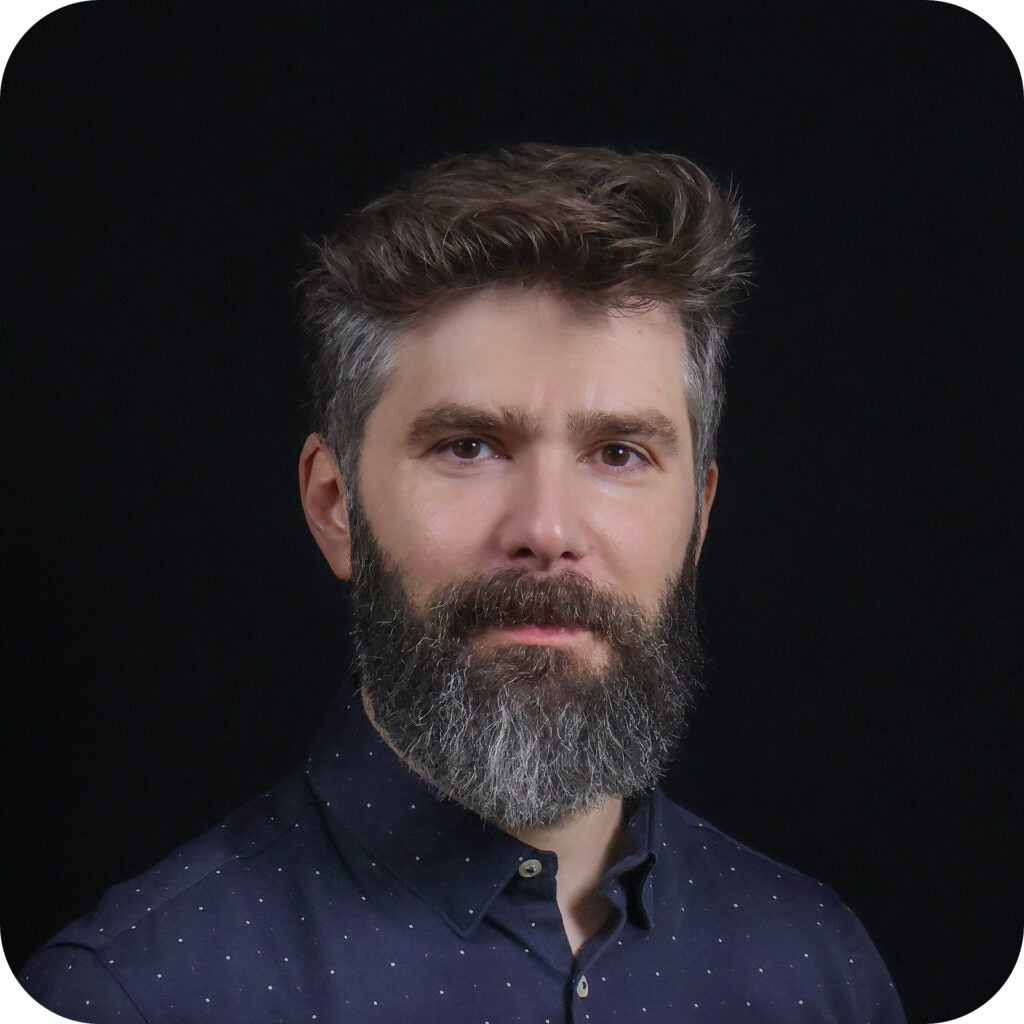 A man with short, wavy brown hair and a full gray beard wearing a dark, collared shirt stands against a plain black background.