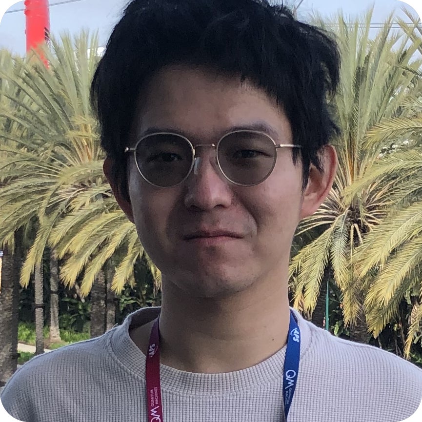 A person with short black hair and glasses stands outdoors in front of palm trees, wearing a light sweater and a lanyard.