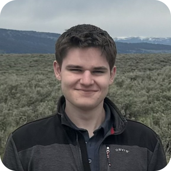 A young man with short brown hair stands outdoors in front of a shrub-filled landscape and distant mountains under a cloudy sky.