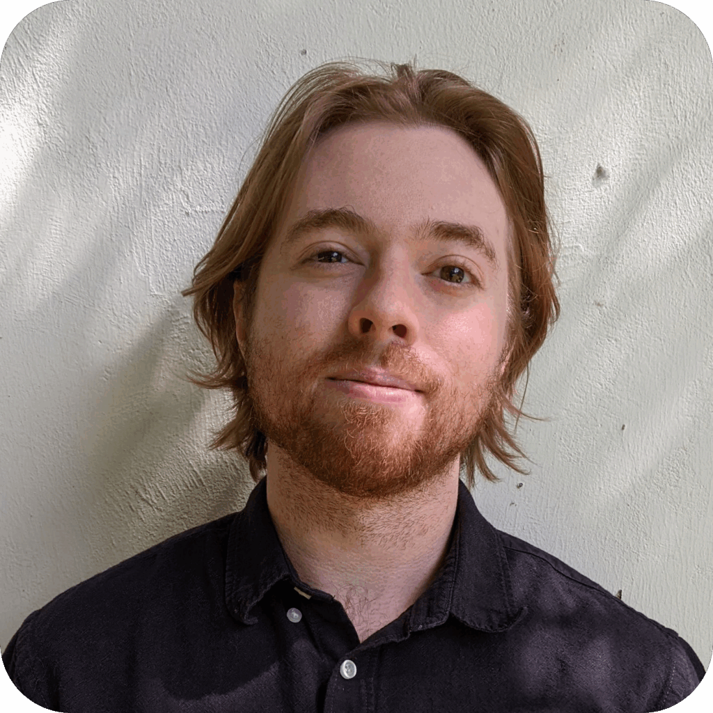 A person with light brown hair and a beard, wearing a black collared shirt, stands in front of a plain white wall.