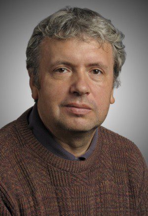 Middle-aged man with curly gray hair, wearing a knitted sweater, against a gray background.