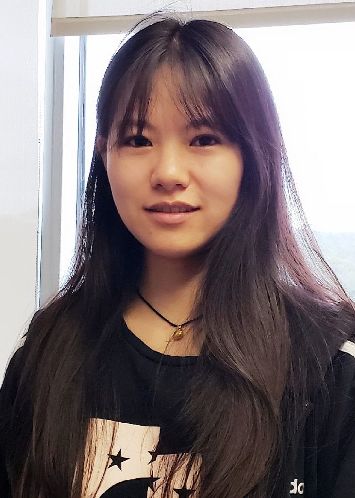 A young woman with long black hair wearing a black t-shirt stands indoors, smiling slightly at the camera.